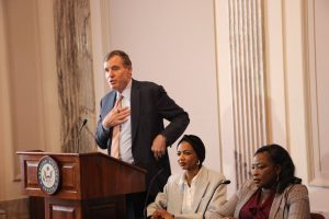 Virginia Senator Mark Warner (D-IN) standing behind a podium, in speech. Niemat Ahmadi with another woman sit to his right at a lower table, with stoic faces, listening.